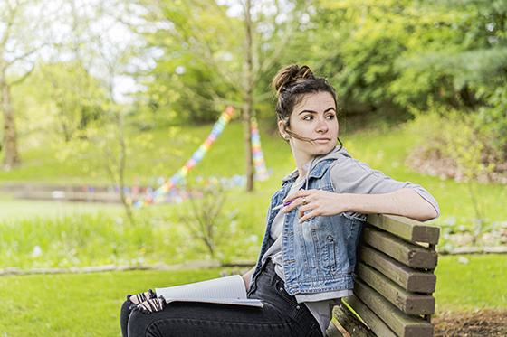 Photo of a Chatham University student sitting on a bench outside writing in notebook on lap. 