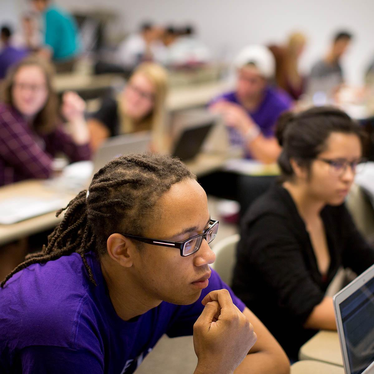 Photo of students listening in a classroom, with a young Black man with dreads, 眼镜, and a purple Chatham shirt in the foreground