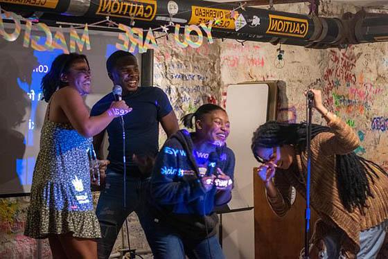 Photo of four 黑色的 students laughing and sining on stage with microphones during BIPOC 卡拉ok night. There is a sign behind them that says YOU ARE MAGIC.